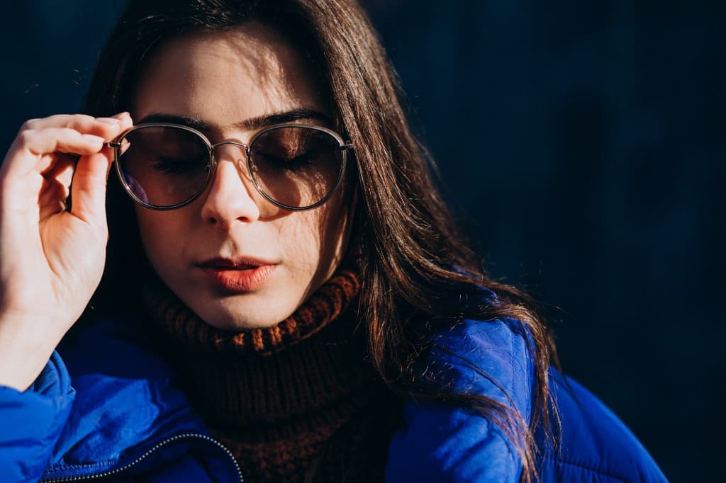 Woman wearing round sunglasses and a blue jacket, touching the frame of her glasses